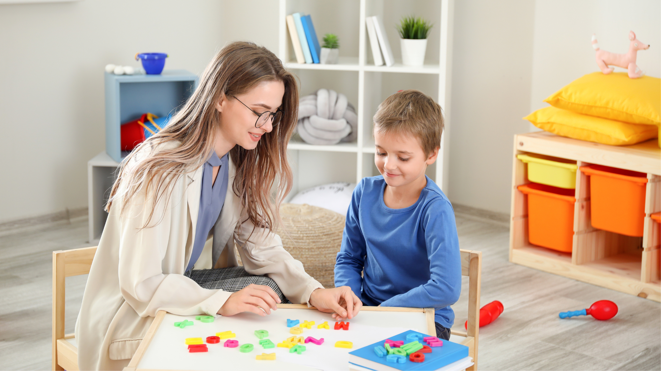 Child therapist sitting at a table with a little boy wearing a blue top looking at magnetic letters on a table in a therapy room