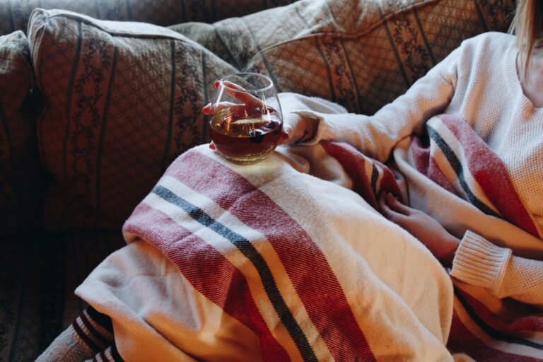 Person on a patterned couch holds a glass of red wine, wrapped in a cozy red-striped blanket.